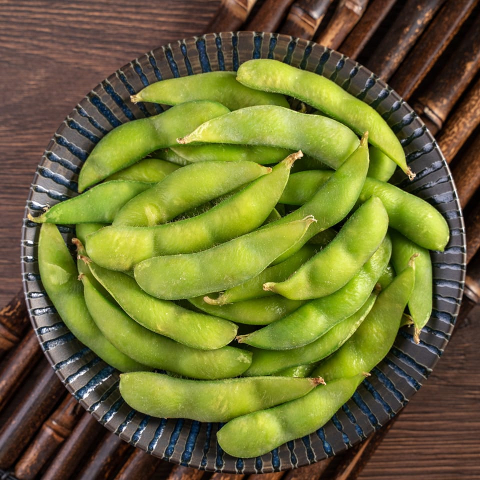 A dark blue and black bowl filled with bright green edamame pods, on a bamboo mat.