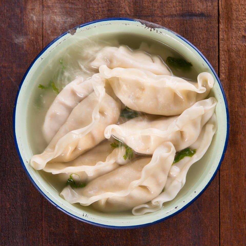 Steaming hot dumplings in a light broth with green herbs, served in a rustic bowl.